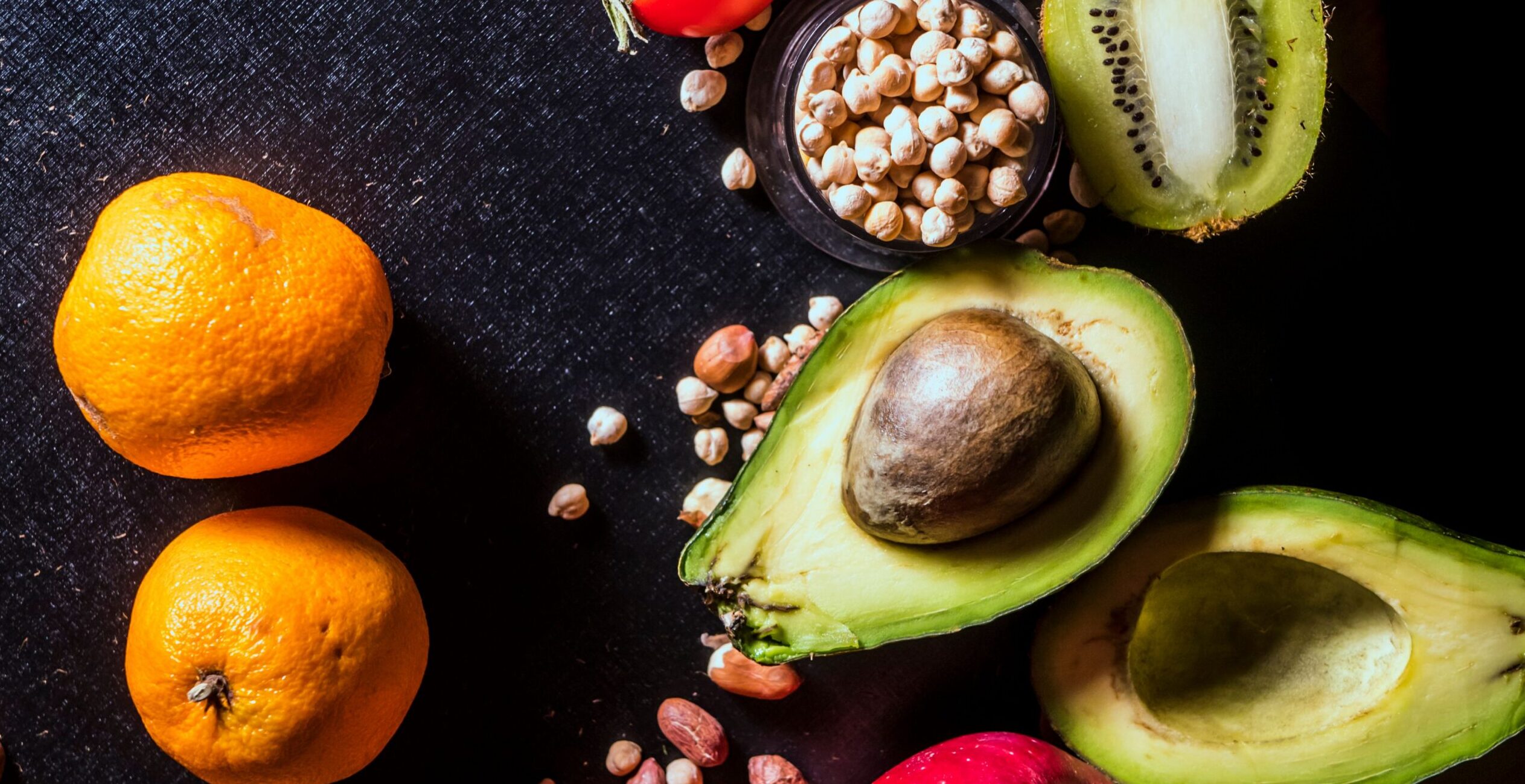 A vibrant arrangement of fruits and nuts on a black background featuring kiwi, avocado, oranges, apple, and peanuts.