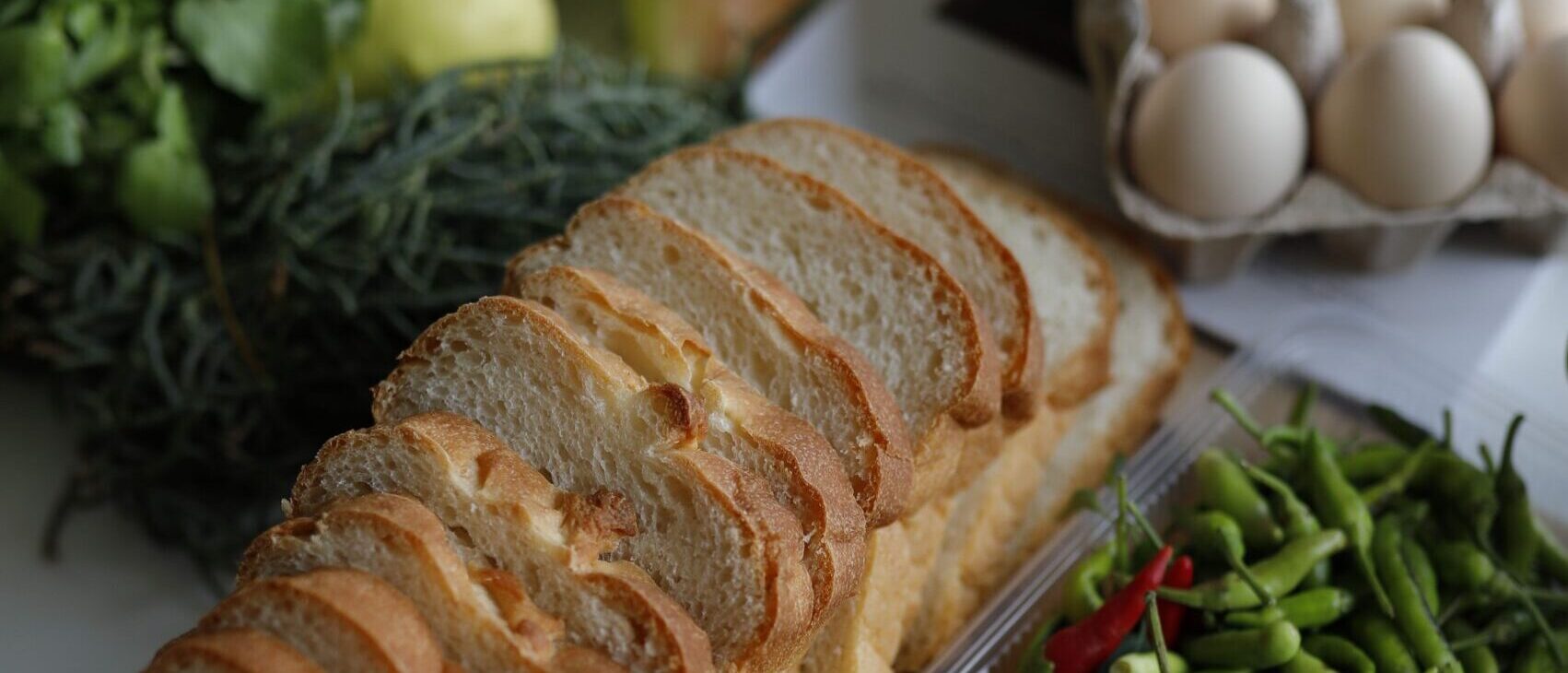 A vertical overhead shot of sliced bread, fresh vegetables, and eggs on a wooden board in a kitchen setting.
